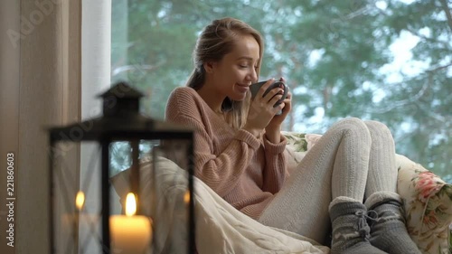 Cozy winter lifestyle. Happy young woman enjoy of cup of hot coffee sitting home by the big window with winter snow tree background