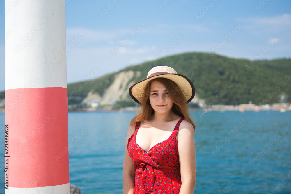 A beautiful young woman in a hat and a red dress walks by the sea. Portrait