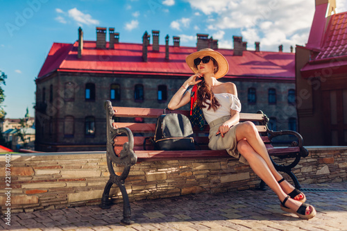Young stylish woman wearing hat and sunglasses outdoors. Stylish traveler girl resting on bench against old architecture