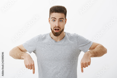 Guy learned how gig economy works being amazed and intrigued posing in grey t-shirt looking at camera shocked with opened mouth pointing down being questioned and surprised over white wall
