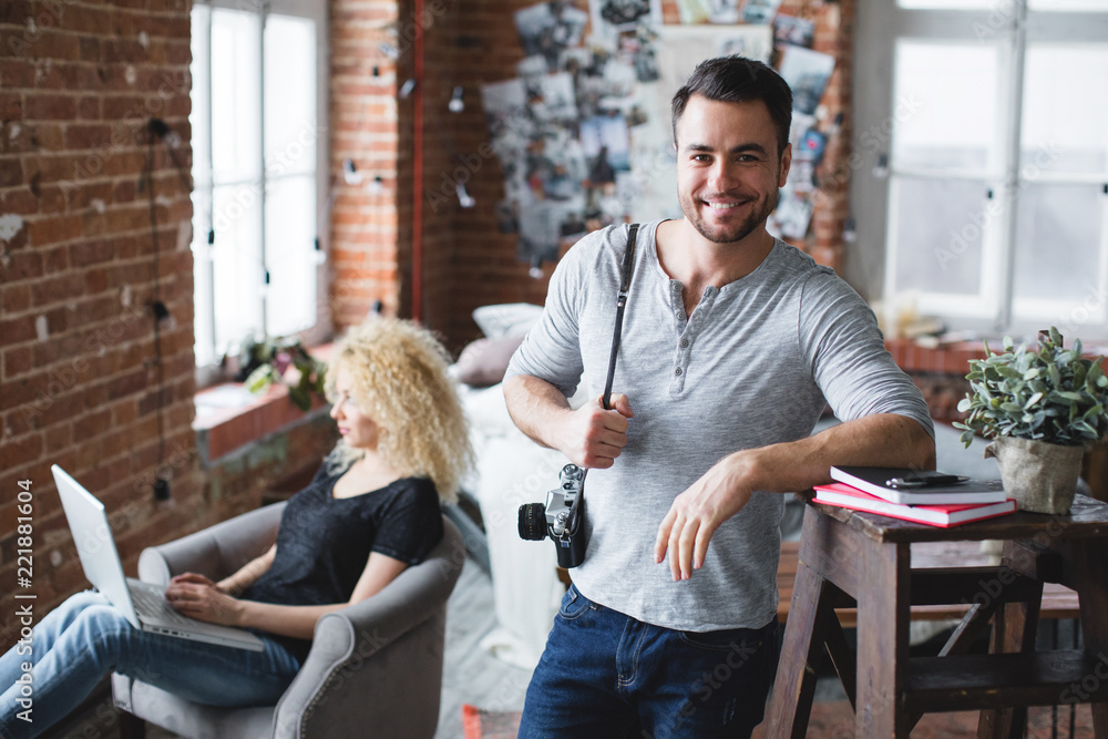 Fototapeta premium Smiling man in a grey t-shirt and jeans standing and a woman sitting with a laptop in loft.