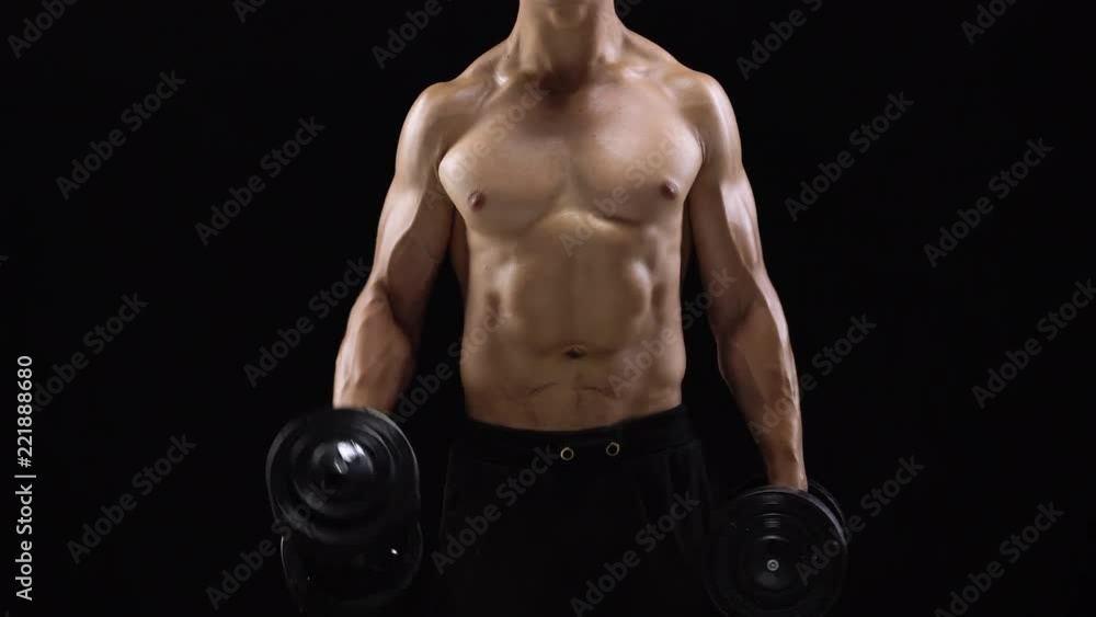 Man flexes his hands with dumbbells, training his biceps on a black background in the studio