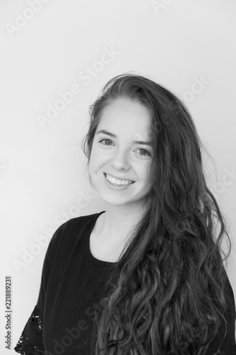 Cute brunette with long hair and black dress looking at camera and smiling with teeth, shoulders turned. White wall background. B&W portrait of cute Caucasian girl with long hair wearing black dress.