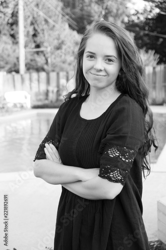 Young woman standing with her arms crossed wearing a black dress in the backyard with a wooden fence and swimming pool in the backyard. 3/4 view, black and white portrait.