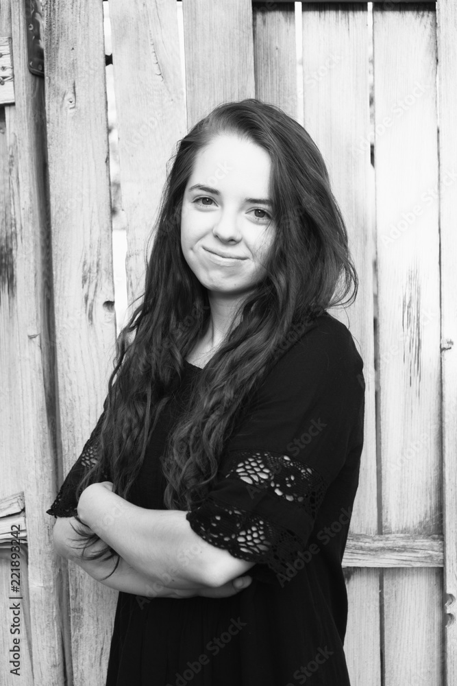 Fototapeta premium Brunette woman posing in front of a rustic old wooden fence wearing a black dress standing with arms folded 3/4 view. Portrait of a young Caucasian woman standing in front of a fence outside, b&w.