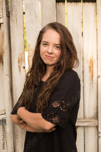 Brunette woman posing in front of a rustic old wooden fence wearing a black dress standing with arms folded 3/4 view. Portrait of a young Caucasian woman standing in front of a fence outside.