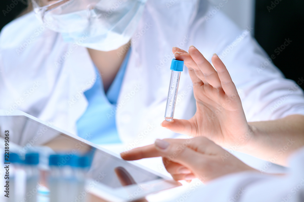 Closeup of scientific research team with clear solution in laboratory. Blonde female chemist holds test tube of glass while her colleague checks results with tablet pc. Blood test, medicine or Closeup of scientific research team with clear solution in laboratory. Blonde female chemist holds test tube of glass while her colleague checks results with tablet pc. Blood test, medicine or