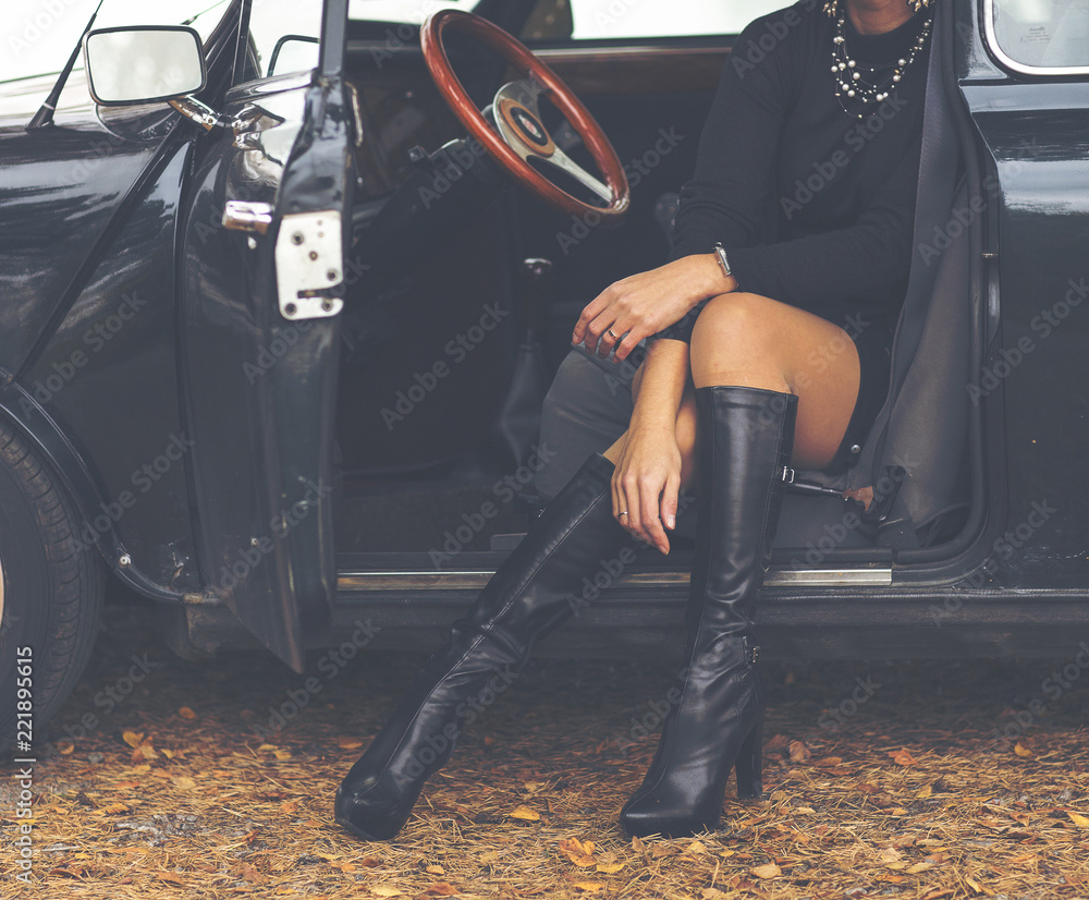 Sexy lady sitting in a classic car. Long leather boots and black dress. Image has a vintage