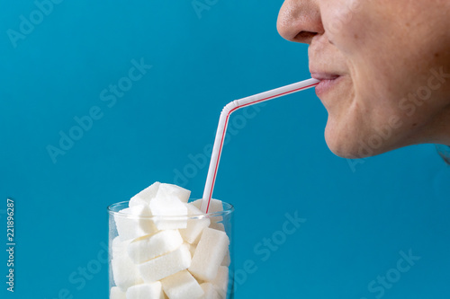 Fotografie Profile of a young woman drinking with a red stripes straw from a glass filled with sugar cubes on blue background