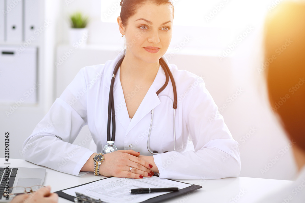 Brunette female doctor talking to patient at hospital office. Physician says about medical exams results for choosing optimal treatment. Healthcare and medicine concept Brunette female doctor talking to patient at hospital office. Physician says about medical exams results for choosing optimal treatment. Healthcare and medicine concept