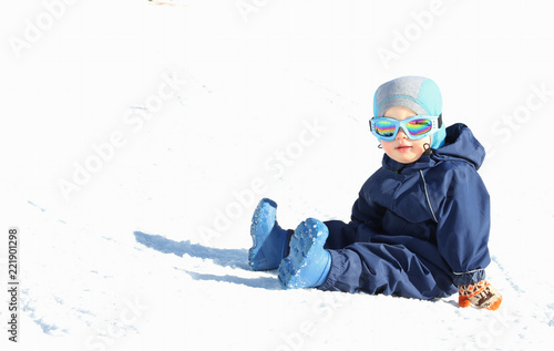 Cute toddler boy in winter suit and ski glusses sits on snow slope on sunny day. Copy space photo