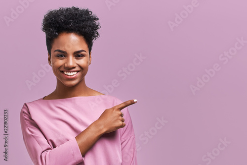 Horizontal portrait of happy dark skinned mixed race female model indicates with fore finger at blank copy space, shows place for your advertisement or promotional text. Lovely African American girl