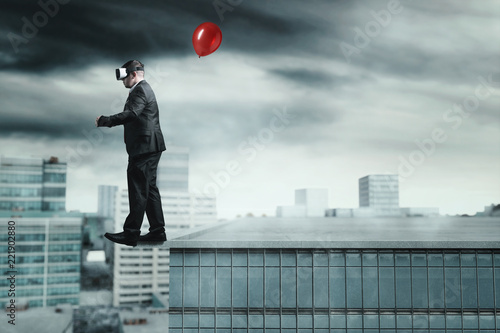Businessman mit VR Brille und Luftballon in der Hand läuft dem Abgrund eines Wolkenkratzers entlang photo
