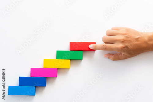 Concept of building success foundation. Women hand put red wooden block on colorful wooden blocks in the shape of a staircase.