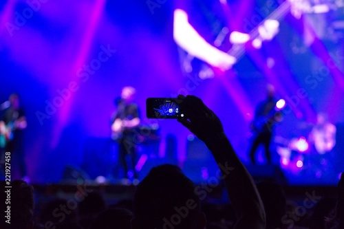 Hand of a man holding a mobile phone and taking a video of a live concert. The background is blurred. blue tone