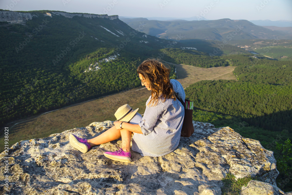 A traveler girl sits on top of a mountain and holds her hat. Active rest concept. Summer view of the mountains with green trees