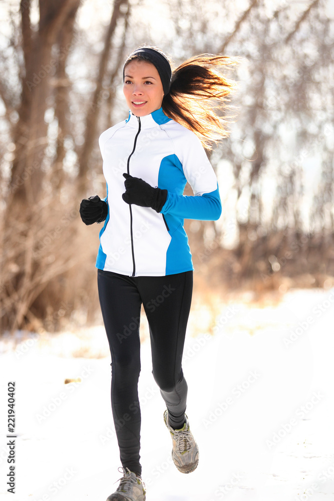 Winter run jogging fit runner girl running outdoor in snow. Asian woman  athlete training outside wearing cold weather clothing jacket, tights,  gloves and headband. Stock Photo
