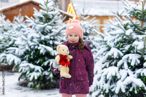 Cute little smiling kid girl on christmas tree market. Happy child in winter clothes and toy choosing xmas tree on xmas market with lights on background on winter snow day.