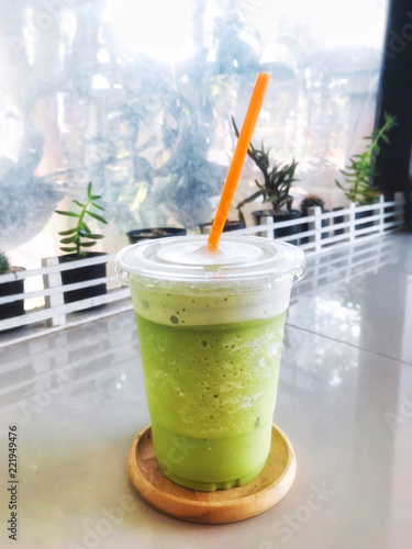 Green tea milk frappe in clear plastic glass have orange straw in wooden tray put on dining table with cactus, succulent and mirror.