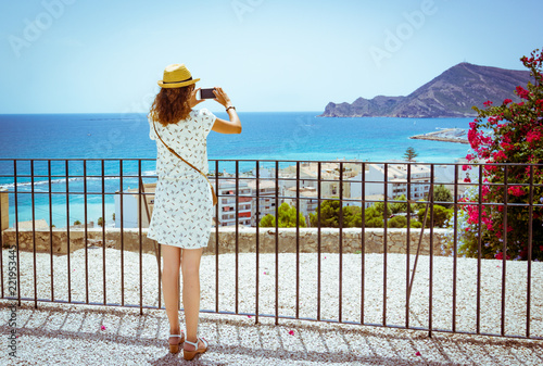 Tourist girl with straw sunhat taking a picture of the mediterranean sea with a smartphone in Altea, Alicante, Spain photo