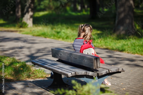 Alone little girl with a backpack sitting on a bench in the Park in the summer