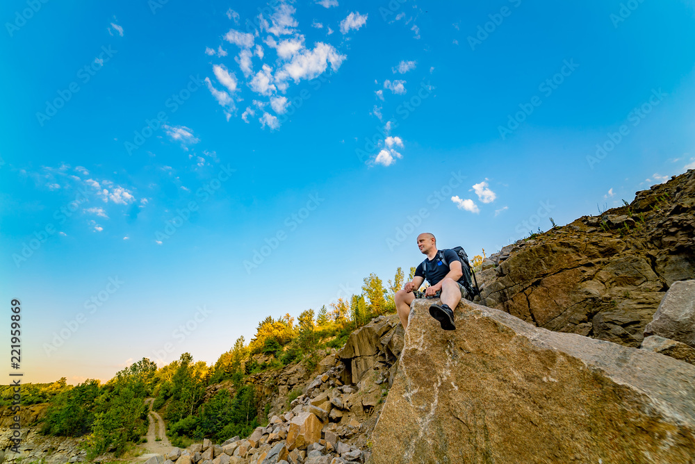 Fototapeta premium a traveler with a backpack sits on a large stone with a sharp end on the background of rock and blue sky in the summer