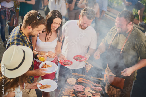Group of people standing around grill  chatting  drinking and eating. 