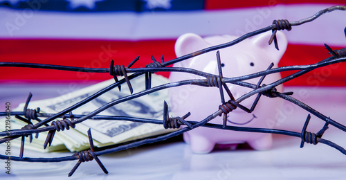 Pink piggy bank and US Dollar money wrapped in barbed wire against United States national flag as symbol of economic warfare, sanctions and embargo busting