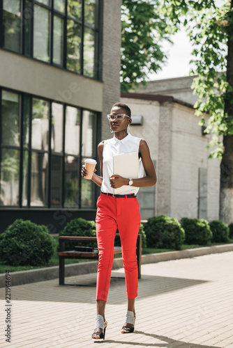 stylish attractive african american businesswoman walking on street with coffee in paper cup and laptop