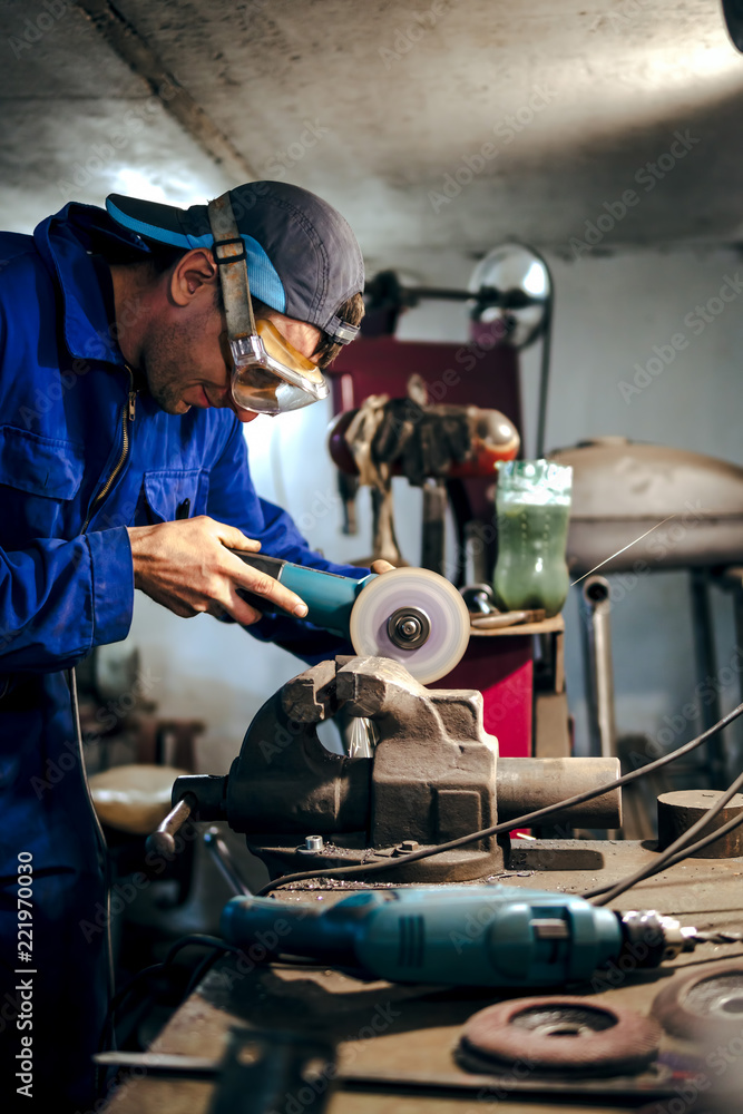 mechanic in blue uniform, cuts the steel part with an angle grinder in the workshop. the concept of protection technology in the workplace, eye protection. sparks fly in the eye, vertical photo