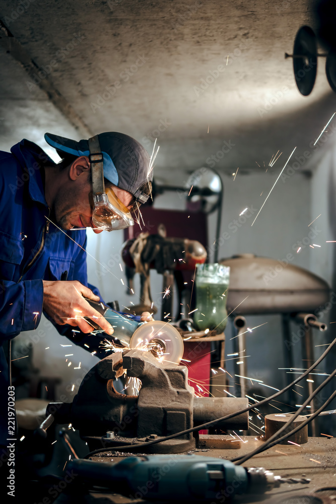 mechanic in blue uniform, cuts the steel part with an angle grinder in the workshop. the concept of protection technology in the workplace, eye protection. sparks fly in the eye, vertical photo