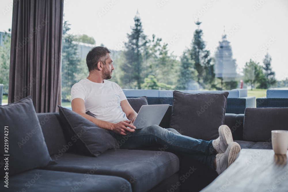 adult male freelancer working with laptop and looking away on sofa at home