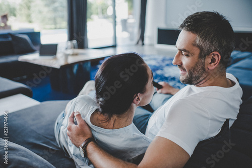 smiling man with remote controller embracing girlfriend and watching tv on sofa at home