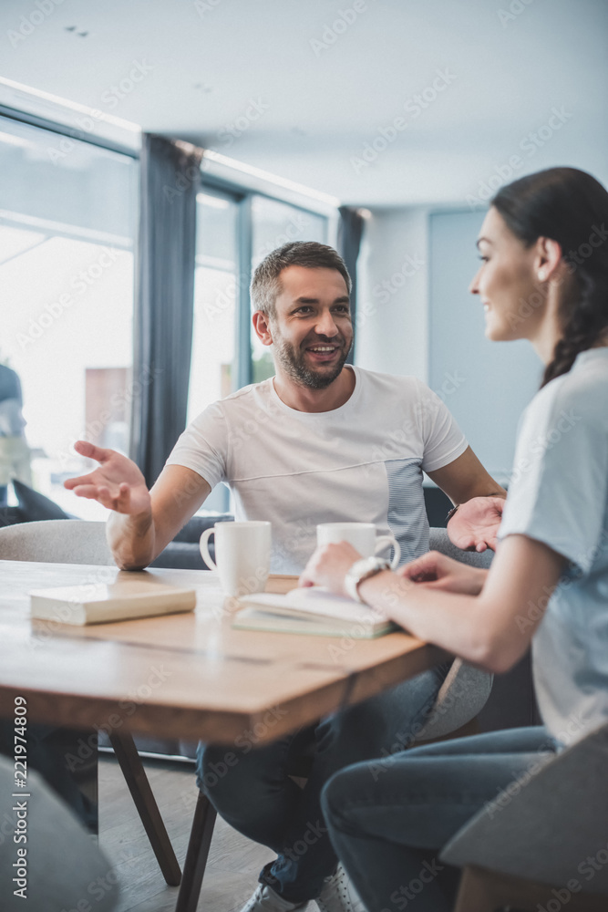 happy adult man gesturing and talking with girlfriend at table with coffee cups and books at home