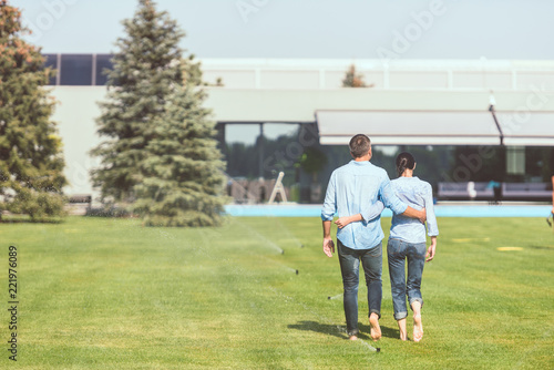rear view of couple embracing each other and walking on lawn near country house