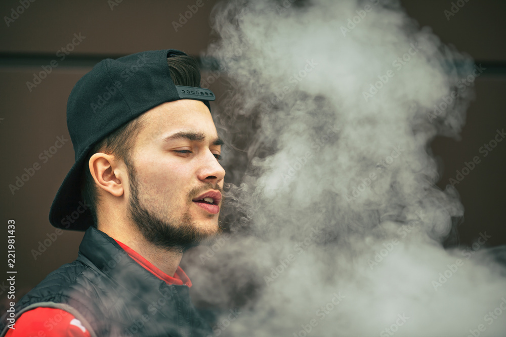Vape teenager. Portrait of a handsome young white man in black cap vaping an electronic cigarette opposite the modern background in autumn.