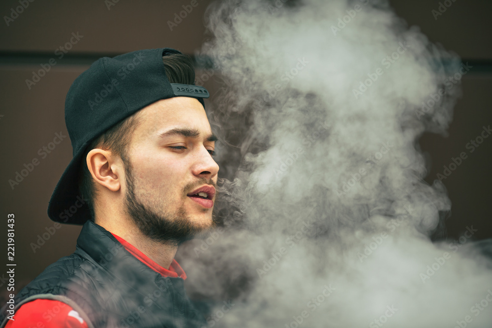 Vape teenager. Portrait of a handsome young white man in black cap vaping an electronic cigarette opposite the modern background in autumn. Vape teenager. Portrait of a handsome young white man in black cap vaping an electronic cigarette opposite the modern background in autumn.