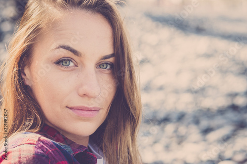 portrait of beautiful young caucasian woman outdoor with stones beach in background. little smile and nice look and blue eyes looking at you. long brown hair and sunlight in background