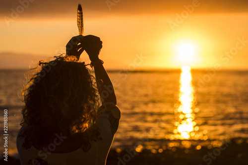 red and oragen colors for dreaming image with girl viewed from rear taking a plume and offer it to the sunset. dream and dreamer concept. ocean and sun in background. travel and vacation image