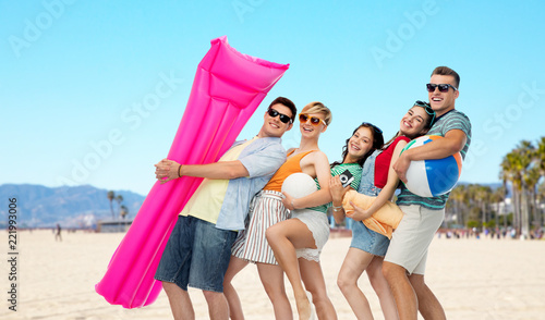 leisure and summer holidays concept - group of happy smiling friends in sunglasses with ball, volleyball, towel, camera and air mattress over venice beach background
