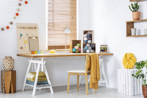 Yellow blanket on chair at wooden desk with lamp and colorful yarns in modern interior. Real photo