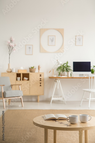 Close-up of a coffee table with a book and two cups with a blurred home office interior in the background. Real photo