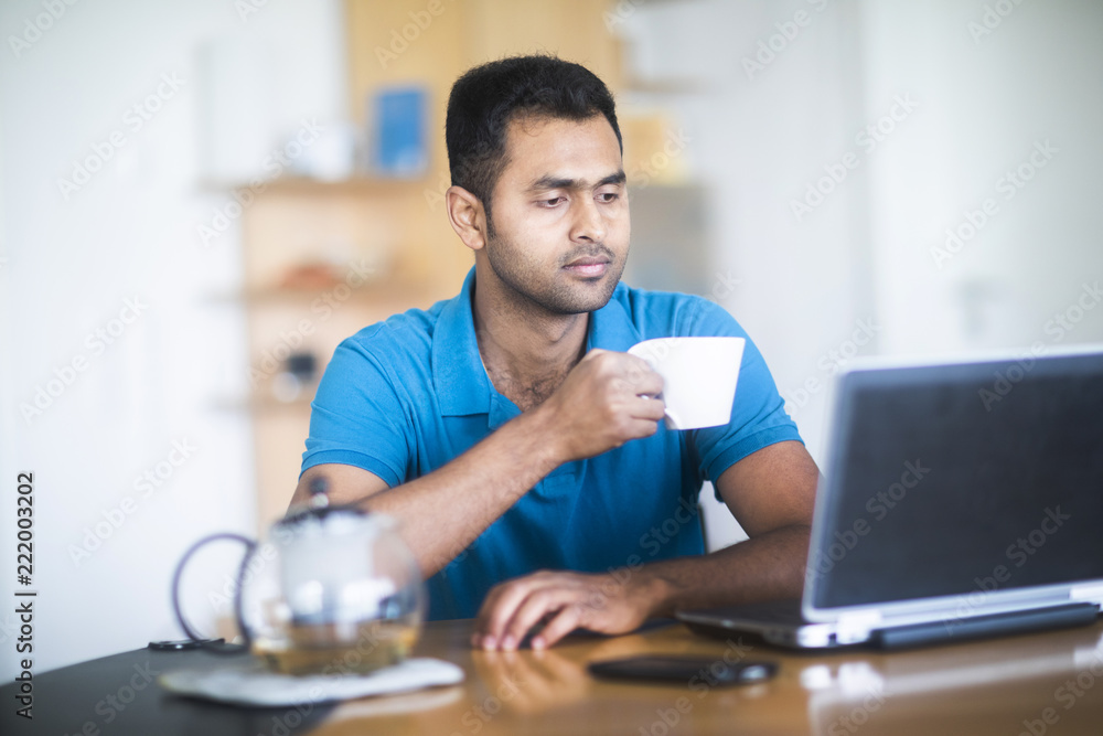 Junger Mann mit Teegefäß arbeitet zu Hause am Schreibtisch Junger Mann mit Teegefäß arbeitet zu Hause am Schreibtisch