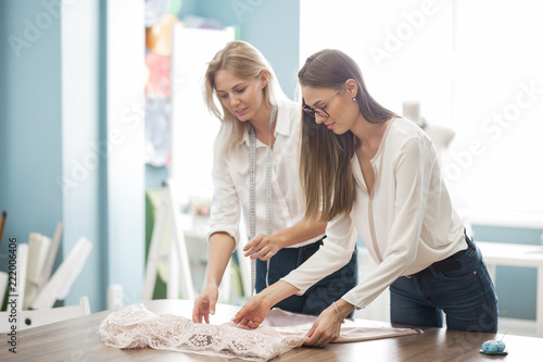 Two smart-looking pretty women wearing white shirts are leaning over the sewing table. Fashion, tailor's workshop.