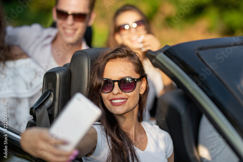 Attractive young brunette woman in sunglasses dressed in a white t-shirt sits with friends in a black cabriolet make a selfie on a sunny day. © Leika production