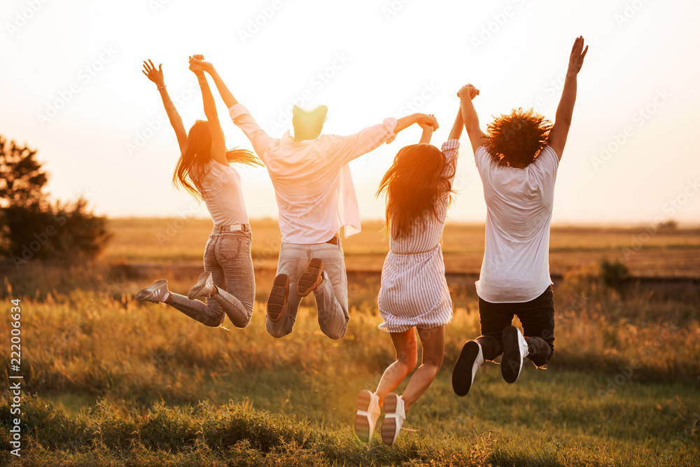 Naklejka premium Dwóch młodych chłopaków i dwie dziewczyny trzymają rękę i skaczą w polu w letni dzień. Widok z tyłu.