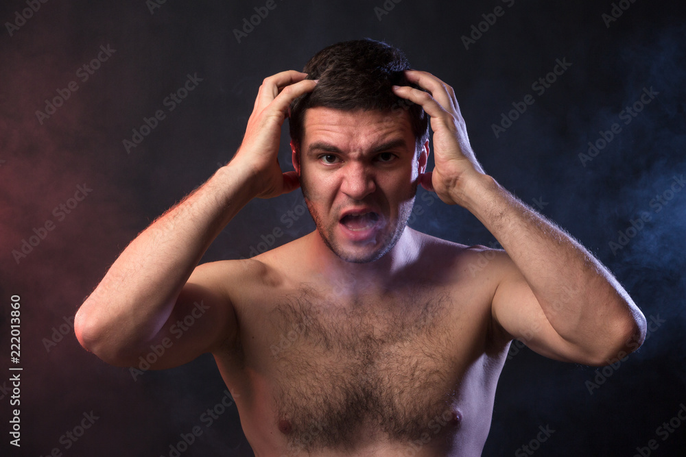 Man grabs his head with his hands and screams from overexertion. Negative emotions of a person. Studio portrait on a black background of a young man of Caucasian appearance. Concept of hysteria Man grabs his head with his hands and screams from overexertion. Negative emotions of a person. Studio portrait on a black background of a young man of Caucasian appearance. Concept of hysteria