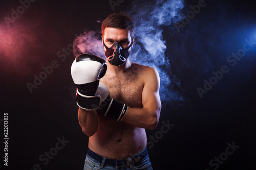 Studio portrait of a muscular boxer in professional gloves of European appearance with light bristles and hair on his chest. Smoke in the background is illuminated in blue and red © Тимур Конев