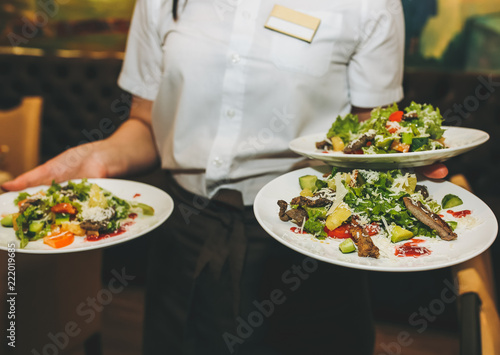 Waitress is holding fresh salad plates in her hand. Woman sets the table at the restaurant. Cafe service for birthday or wedding celebration. Different dishes on the served table.