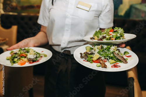 Fototapeta Naklejka Na Ścianę i Meble -  Waitress is holding fresh salad plates in her hand. Woman sets the table at the restaurant. Cafe service for birthday or wedding celebration. Different dishes on the served table.